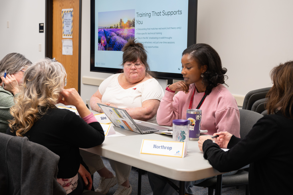 Women sit at a table with cups and a laptop on the table. Behind them is a screen that reads, "Training That Supports You."