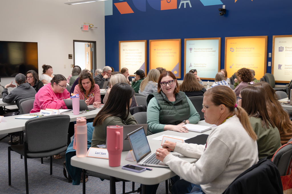 In a room of group discussions at white tables, a woman in a green vest and glasses leans over to make a comment to a peer.