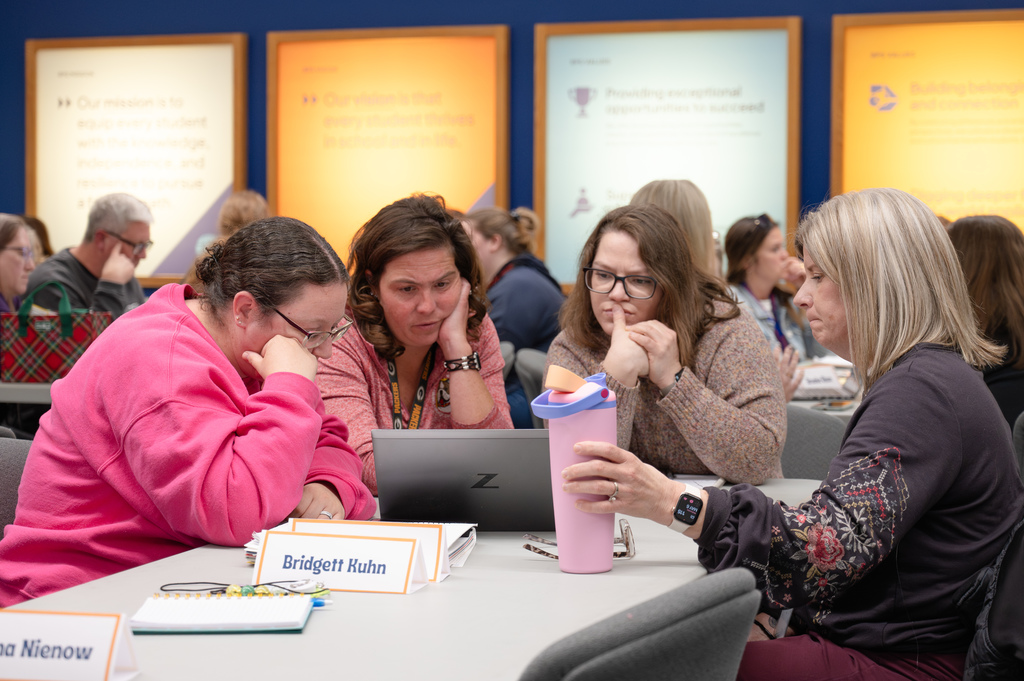 Three women sit and look intently at a laptop. Behind them are lit up posters of the district's values.