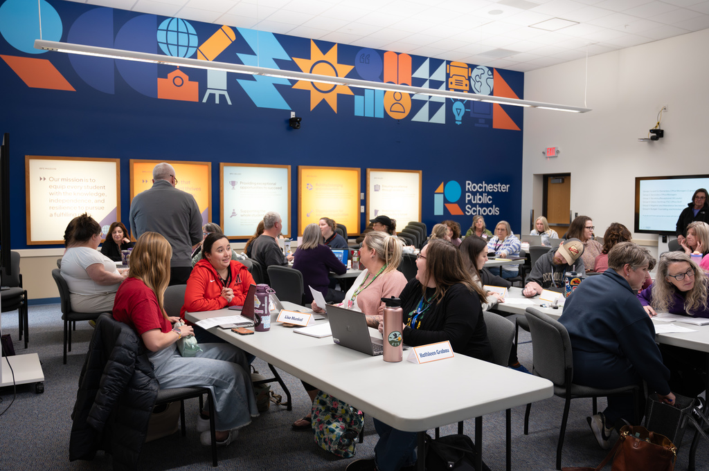 In a large room office managers sit at tables an discuss how to write job descriptions. There are colorful posters on the back wall.