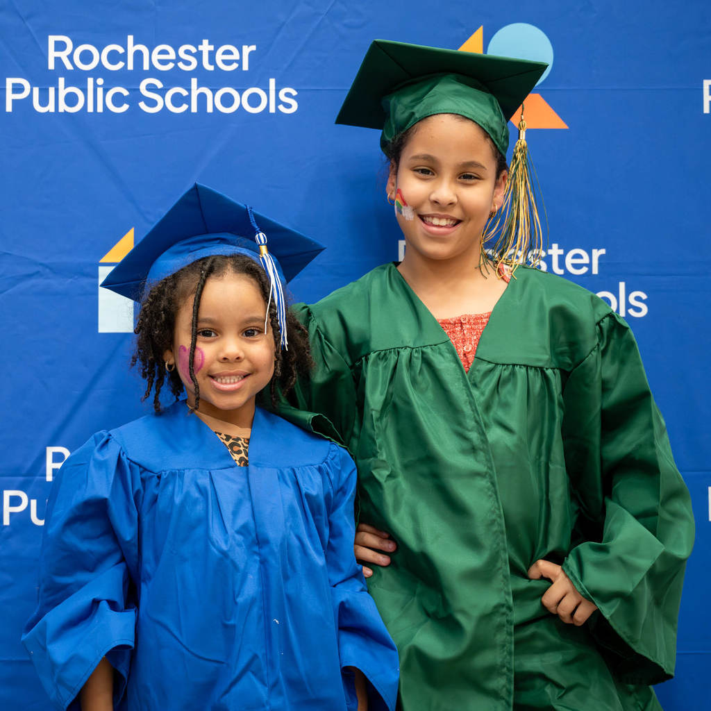 Two sisters dressed in graduation outfits pose in from of a Rochester Public Schools branded background.
