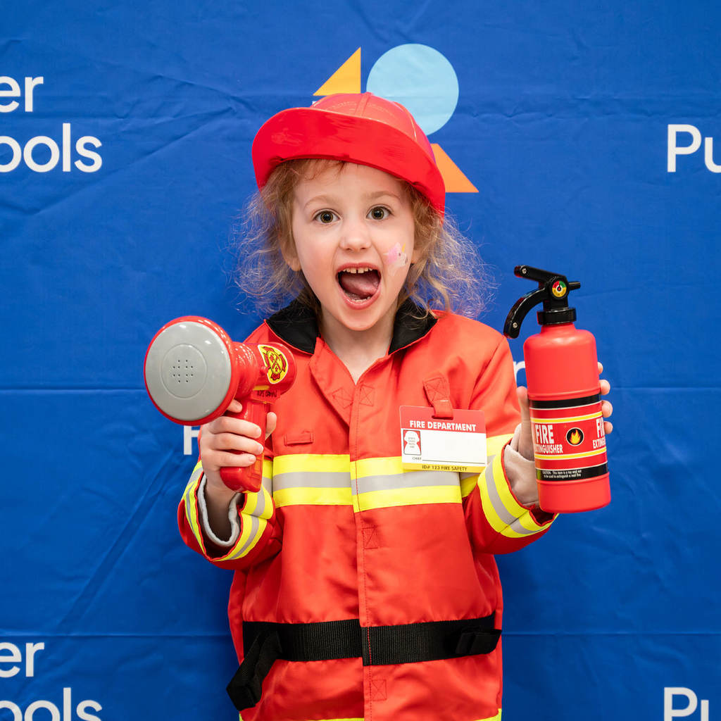 A girl in a firefighter's costume sticks her tongue out in from of a Rochester Public Schools branded background.