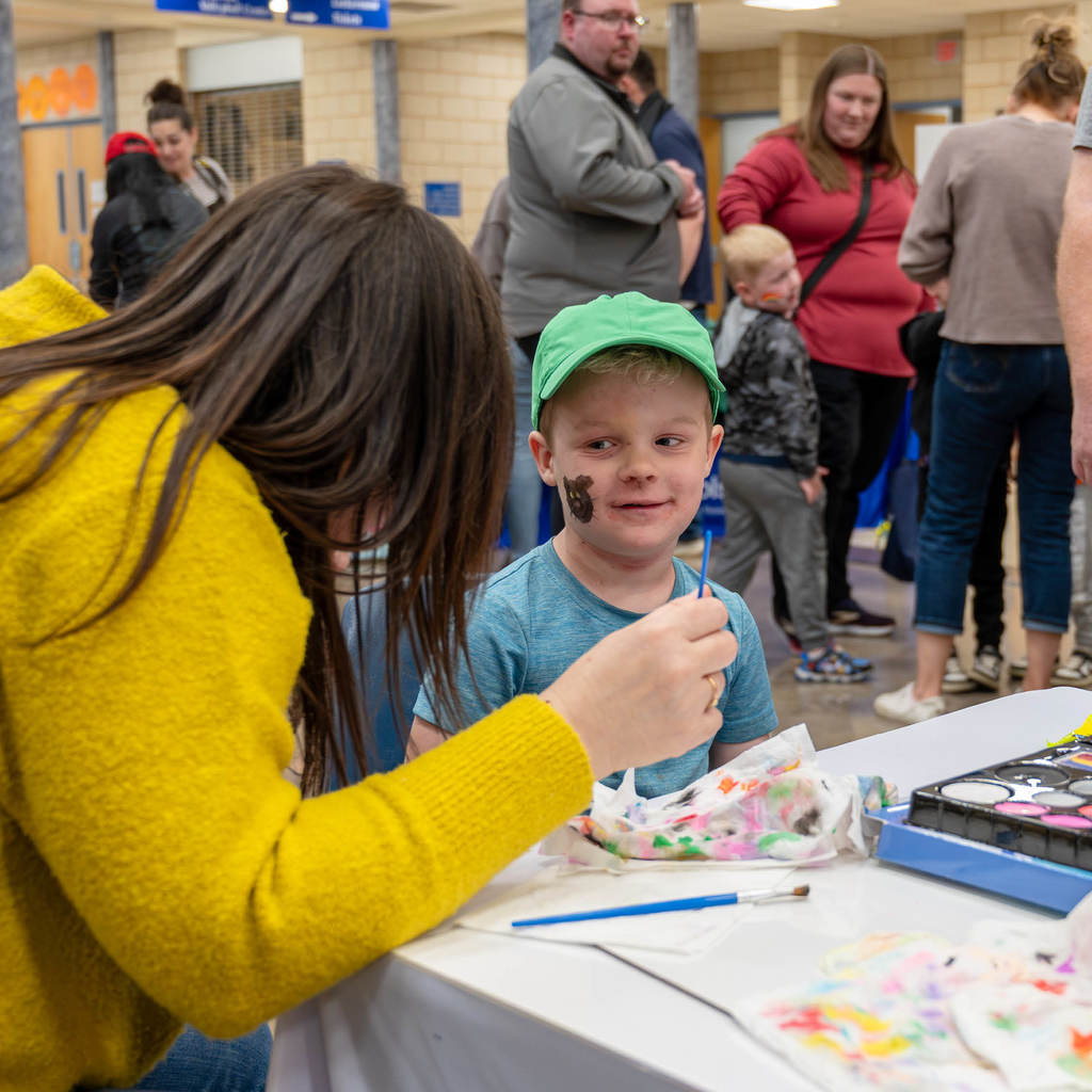 A woman paints a cat on a little boy's cheek. Others mingle in the background.