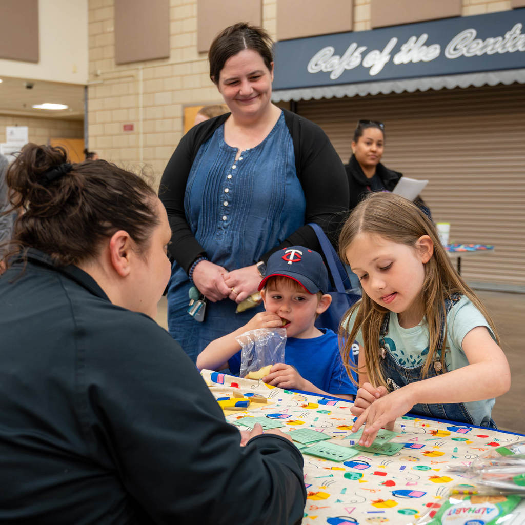 At a booth, a girl focuses intently on matching cards while her young brother eats apple slices and their mom looks on smiling.