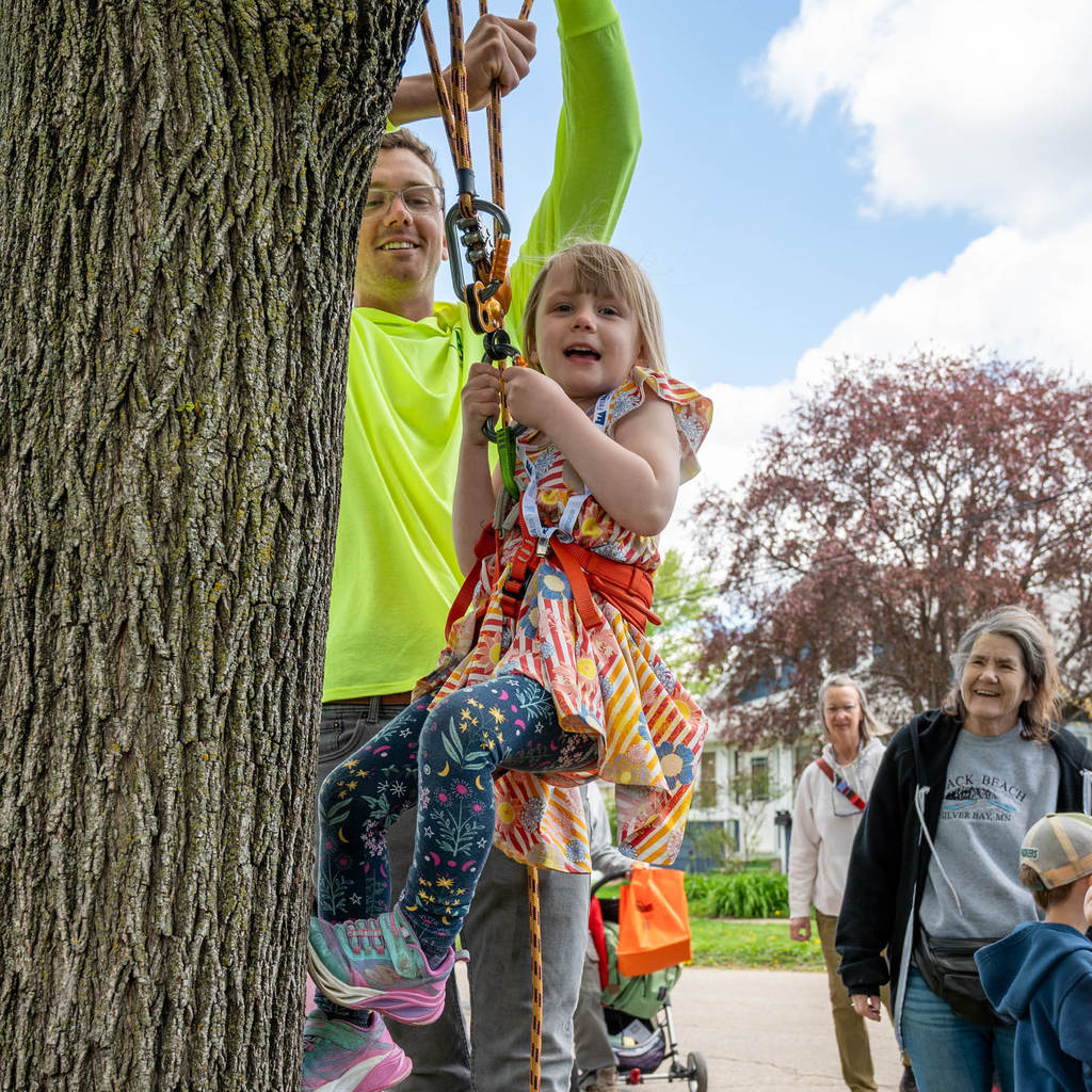 A man in a neon shirt assists a little girl in climbing a few steps up a tree in a harness.