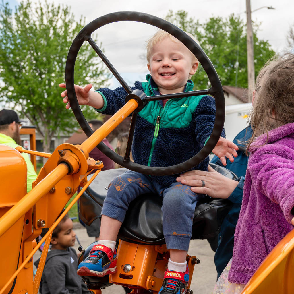 Toddler sits atop a parked tractor and grins as he reaches for the big wheel.