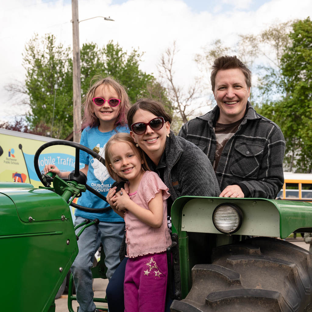 A young family of four sit atop a green tractor and smile at the camera.