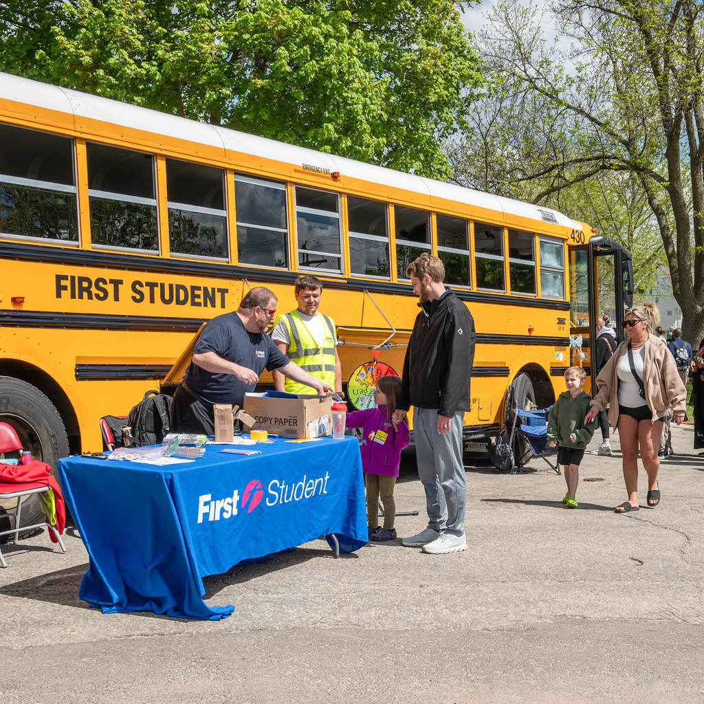 In front of a parked school bus sits a table labeled "First Student." The man at the booth gives a child a handout. Another pair is walking up to the table.