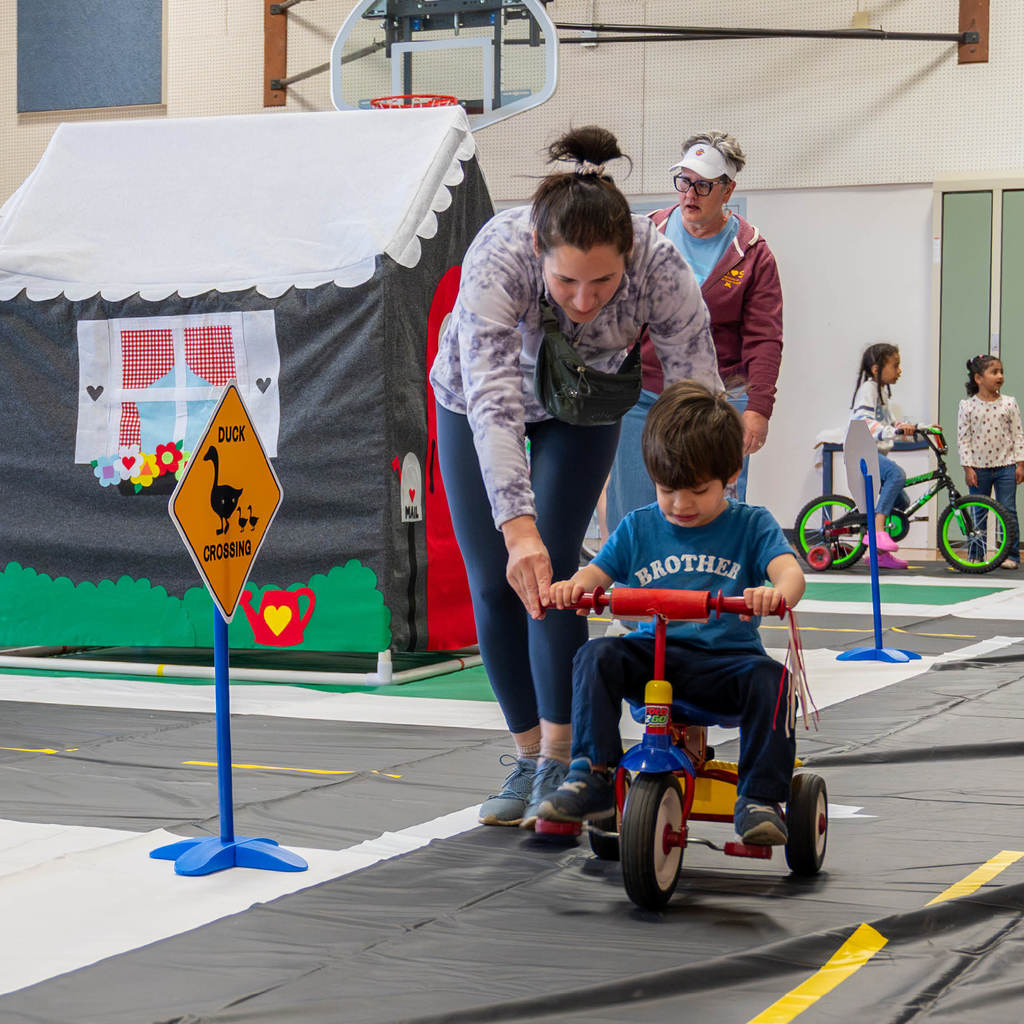 In a gym set up like a neighborhood with roads, an adult helps a toddler ride a tricycle.