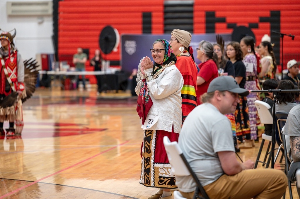 Woman dressed in regalia dancing along to the music on the side of the gym.