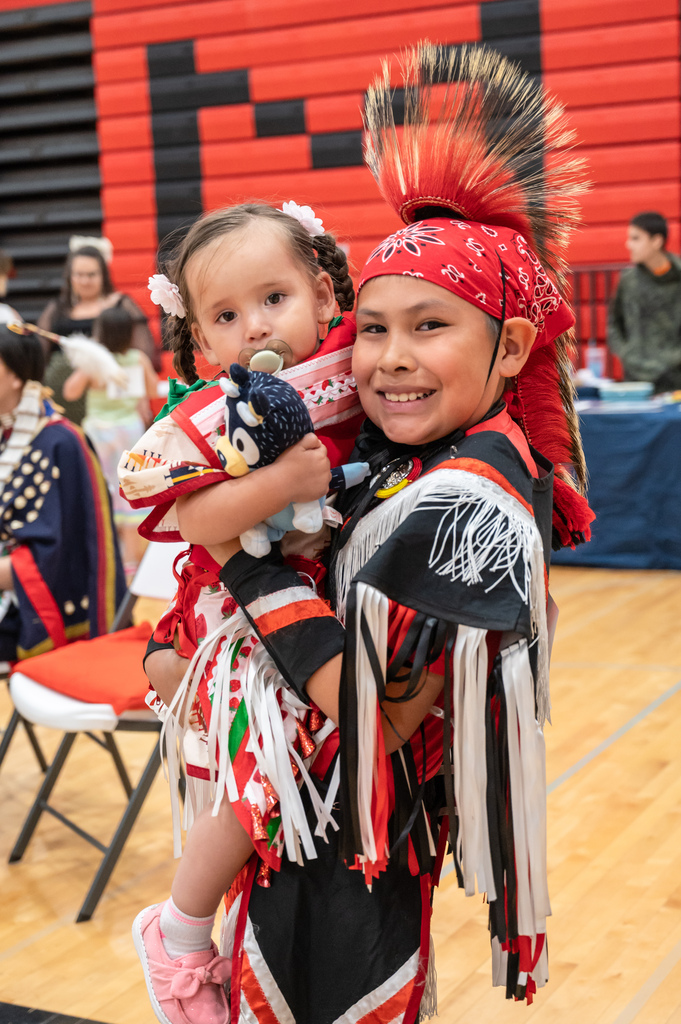 Two young children dressed in regalia hugging and smiling at the camera.