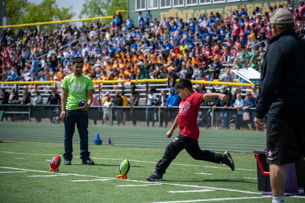 Student leaning back to kick a football with the bleachers full of students in the background.