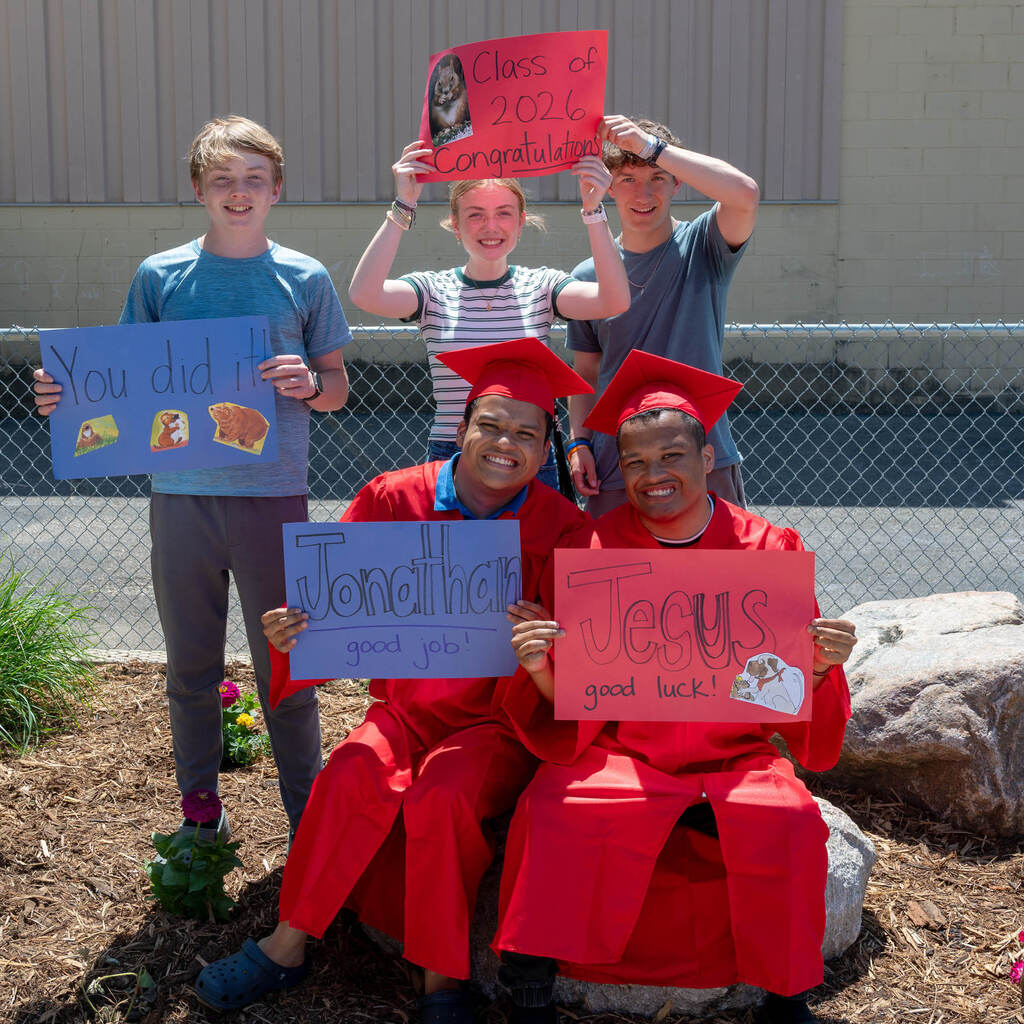 In a posed photo, two grads in red attire hold paper signs that say, "Jonathan good job!" and, "Jesus good job!" Behind them three other students hold paper signs that say "You did it!" and "Class of 2026 congratulations!"