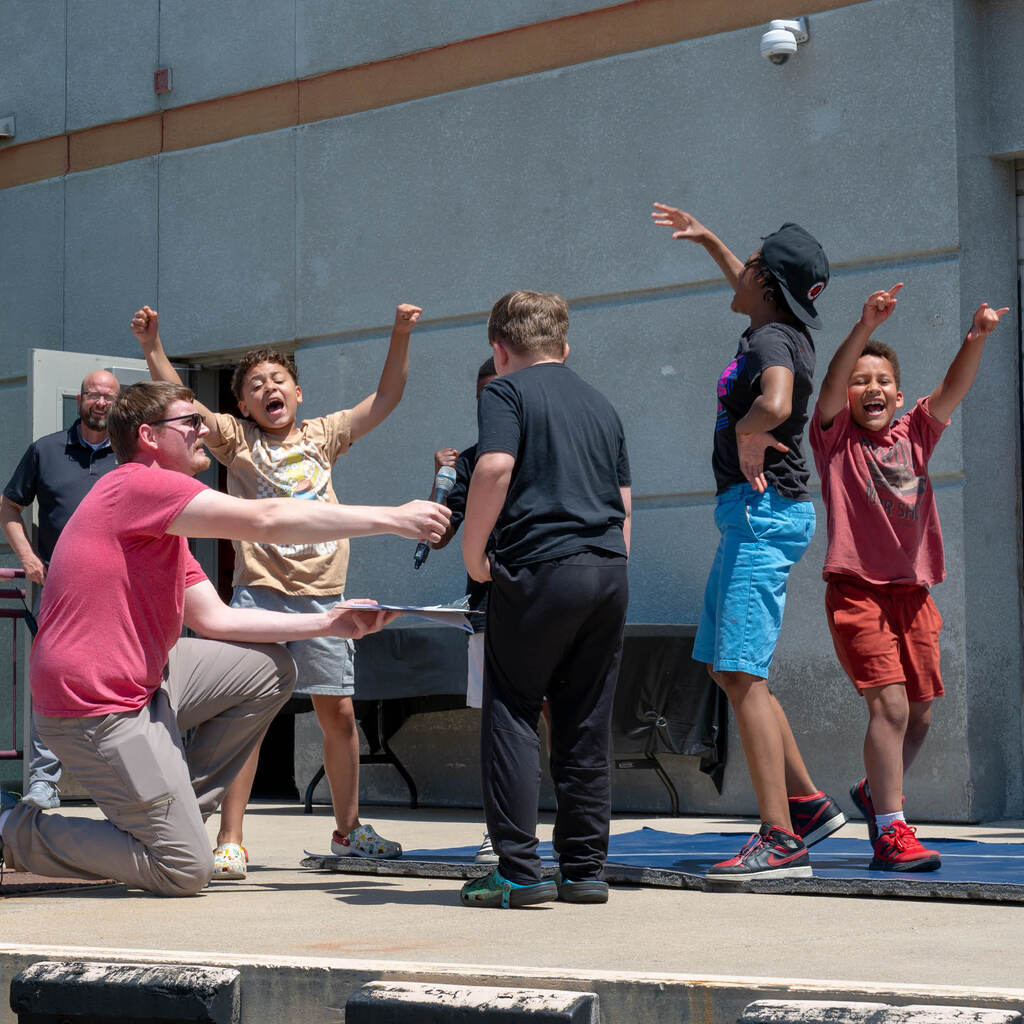 On an outdoor stage, a handful of elementary students dance energetically and rap into a microphone held by a teacher.