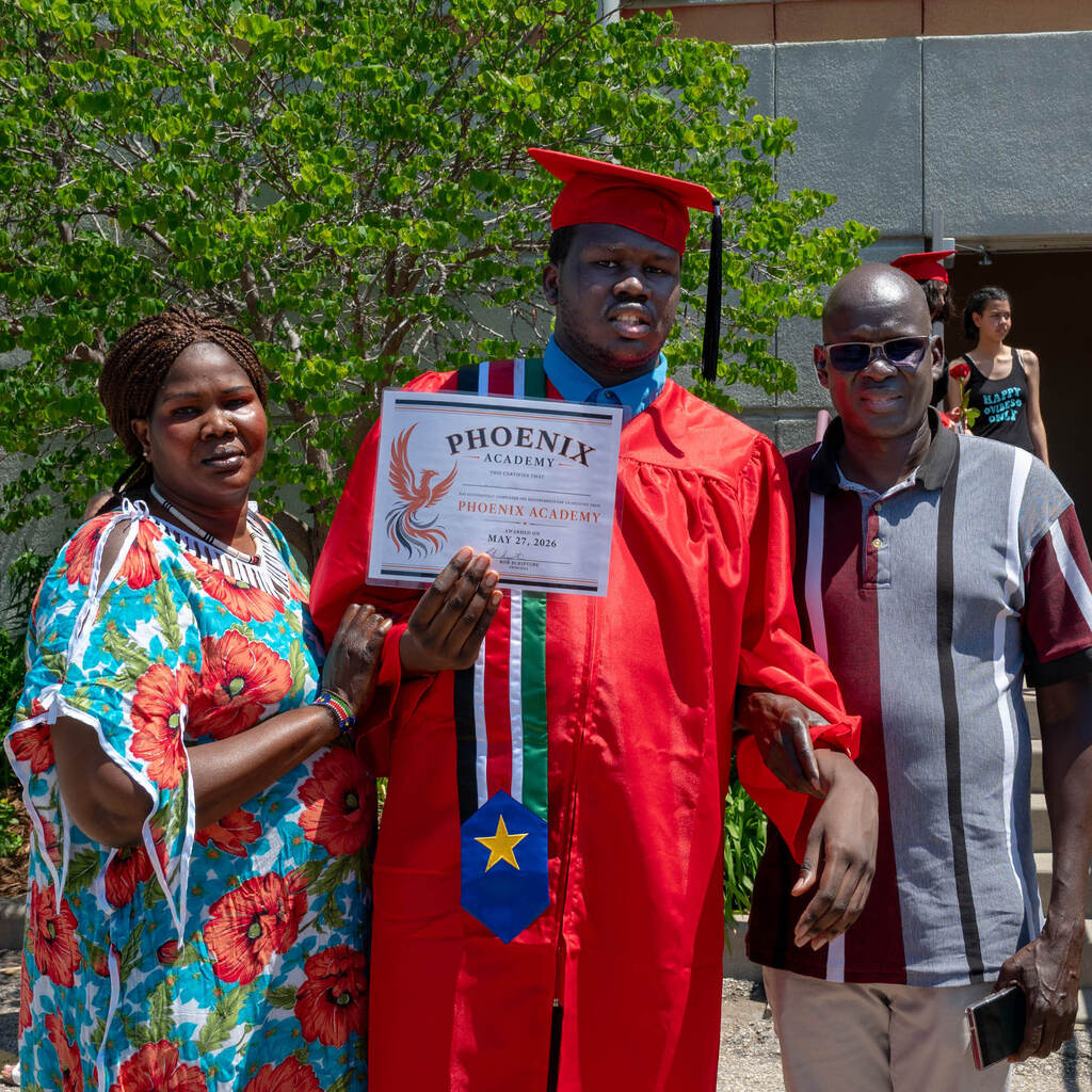 A graduate holds up a diploma that reads, "Phoenix Academy." He stands next to his parents.