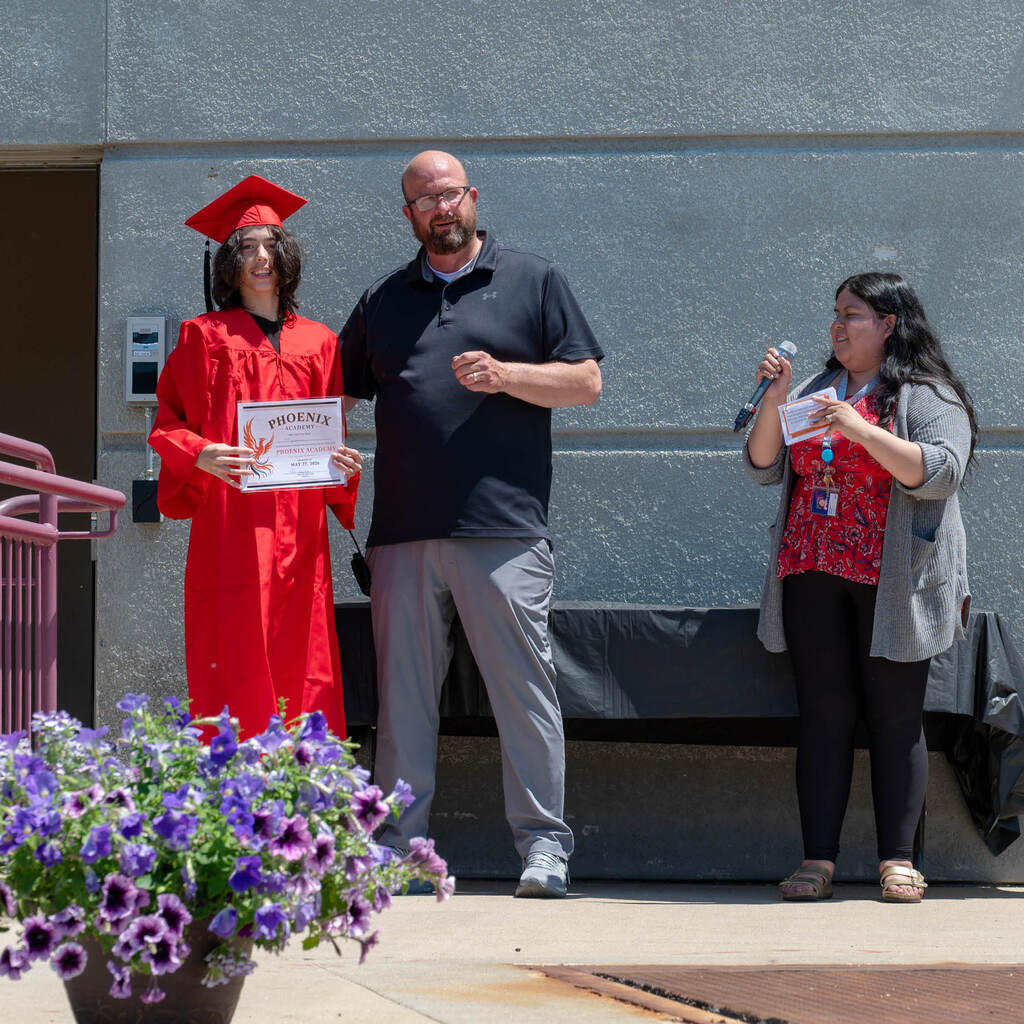 On stage outside, a graduate holds his diploma alongside his principal and a woman with a mic.