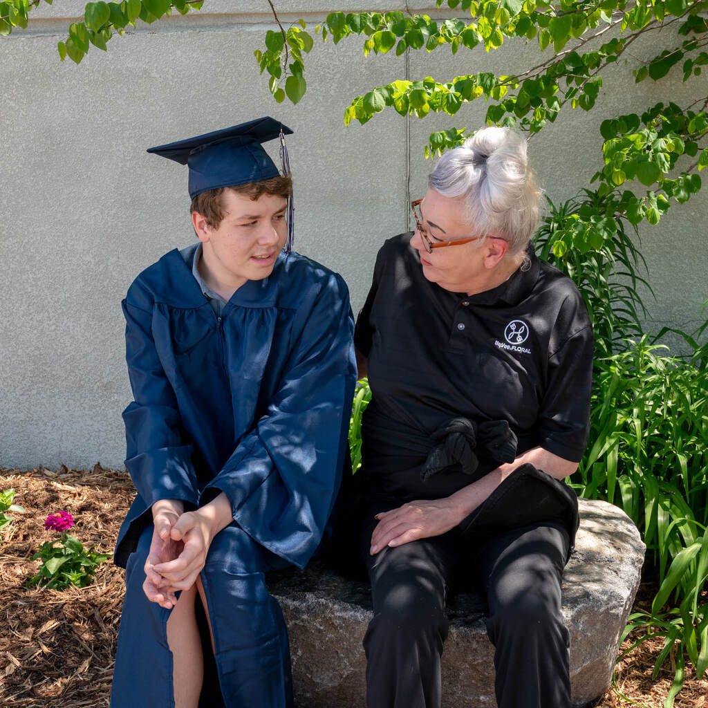 A graduate in a blue robe and cap sits on a landscaped rock with a woman with white hair.