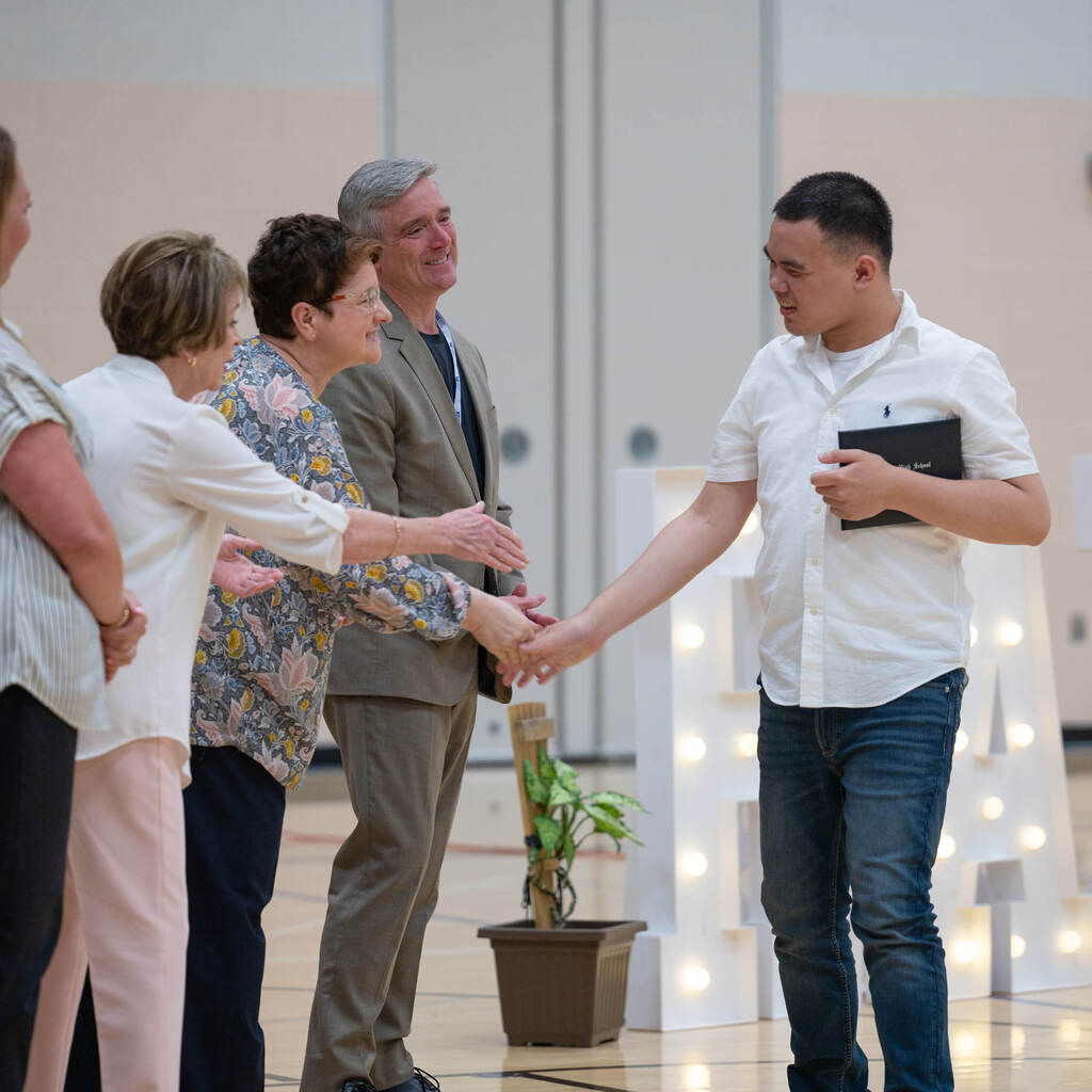 A graduate in a white shirt holds his diploma and shakes the hands of school board members.