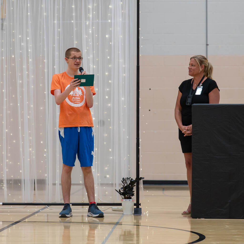 A tall graduate in an orange camp shirt reads an inspirational quote into a microphone while the principal stands off to the side.