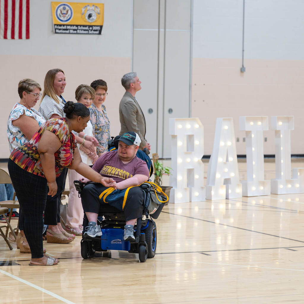 A graduate in a wheelchair rolls along a line of school staff and shakes each of their hands. In the background are giant marquee letters that say, "RAIL."