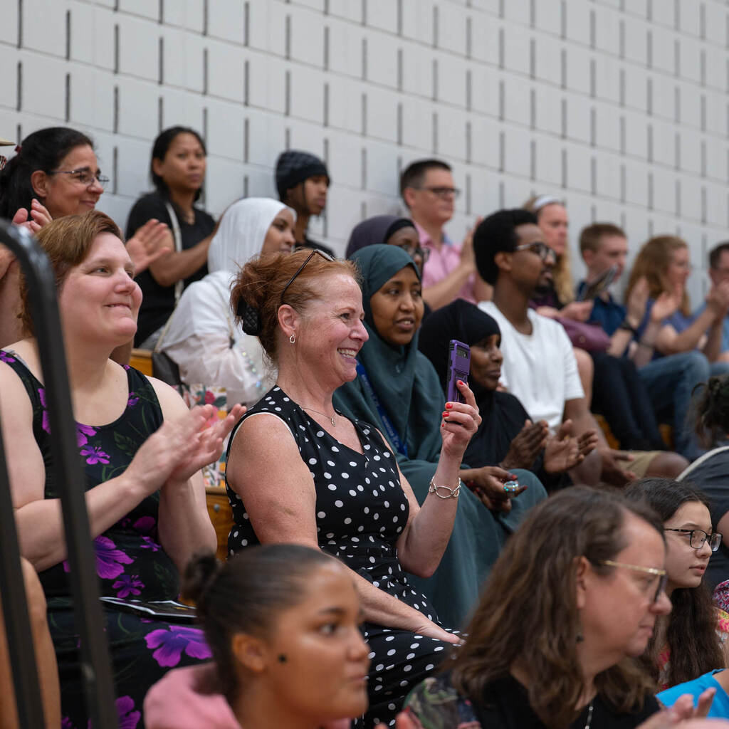 Parents and friends sit in the stands and clap for graduates. The woman in focus grins ear-to-ear and holds up her phone to record the ceremony.