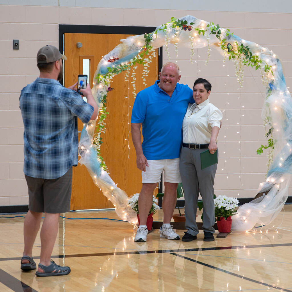 A man takes a picture of a teacher and graduate in front of a lighted arch.