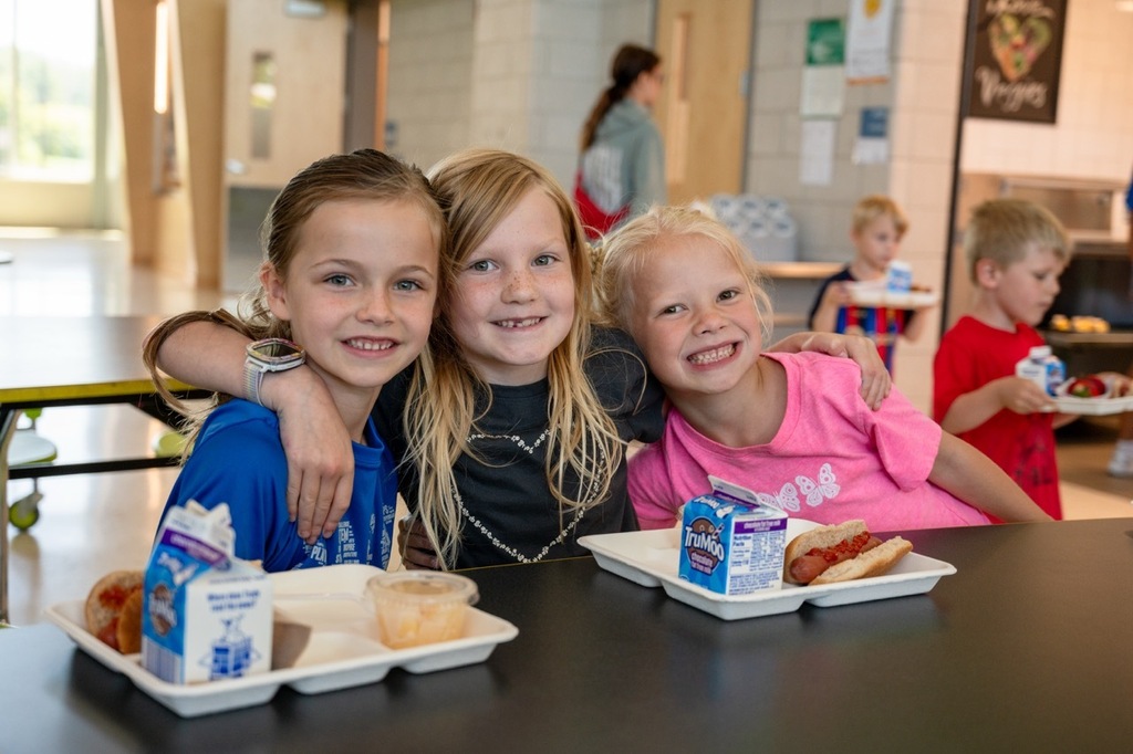 Three girls smiling together sitting at a table with their summer meal lunches sitting in front of them.