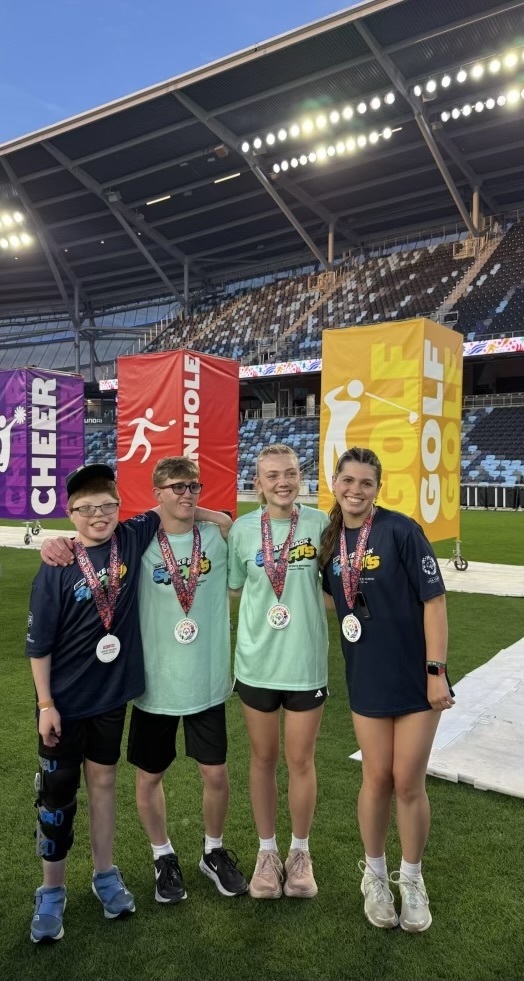 Mayo students arm in arm with their medals in front of Special Olympic signs stating cheer, cornhole, golf.