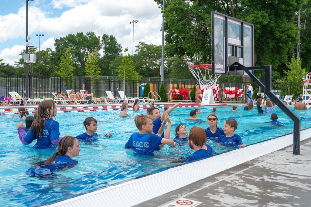 A large group of students in the large pool shooting hoops playing water basketball.