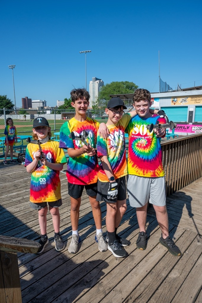 Four students standing arm in arm with their hands on their trash grabbers pointed at the camera and smiling for a photo on the deck of Mayo Stadium.