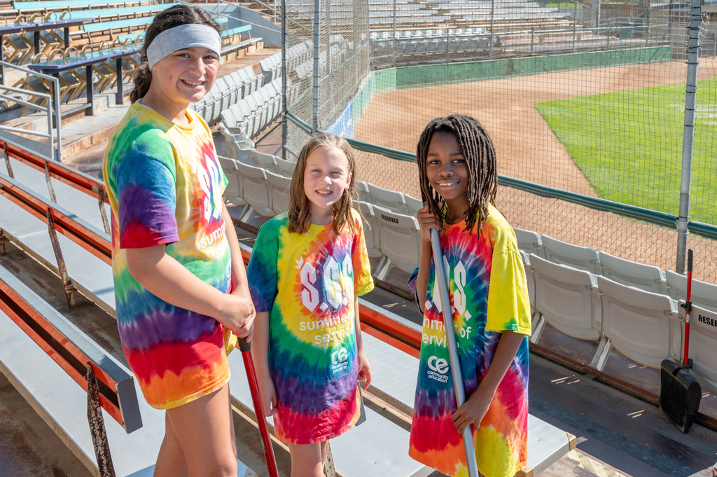 Three students in tie dye sos shirts smiling at the camera with brooms in their hands in the bleachers of Mayo Honker Stadium.