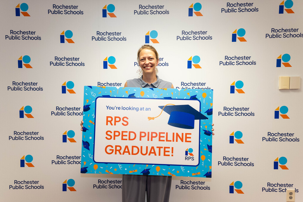 Woman in a gray shirt holds a sign in from of a Rochester Public Schools branded background. Sign reads, "You're looking at an RPS SPED Pipeline Graduate!"