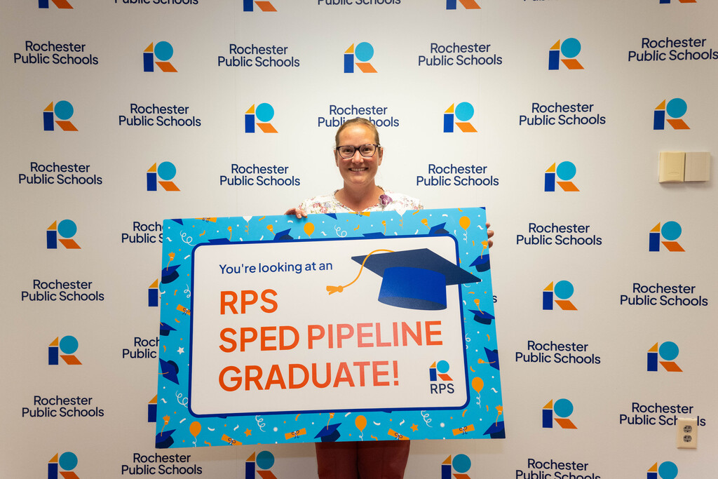 Woman in glasses holds a sign in from of a Rochester Public Schools branded background. Sign reads, "You're looking at an RPS SPED Pipeline Graduate!"