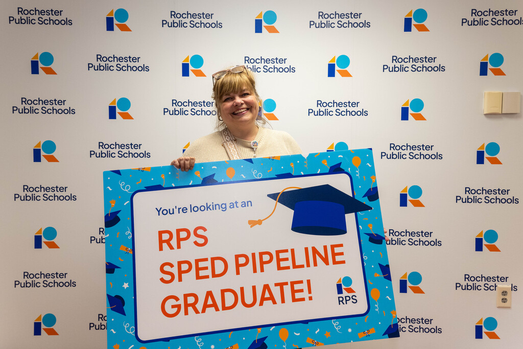 Woman holds a sign in from of a Rochester Public Schools branded background. Sign reads, "You're looking at an RPS SPED Pipeline Graduate!"