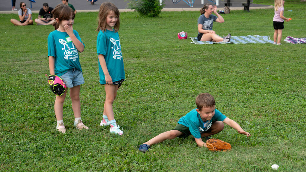 Kids practice retrieving grounders while parents sit on the grass and watch.