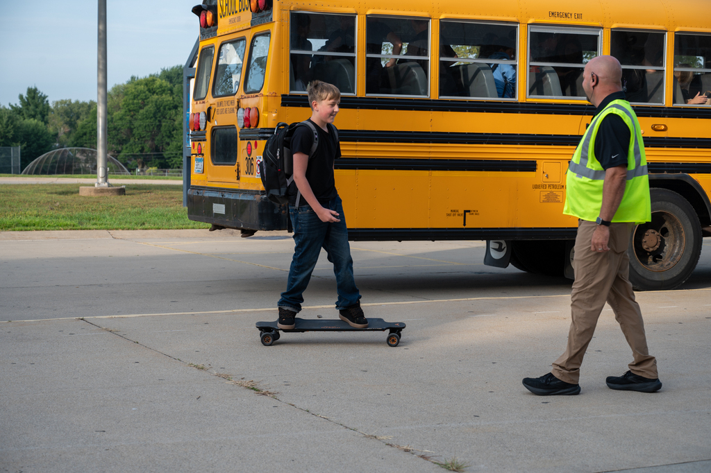 A Kellogg student skateboarding to school and saying hi to Principal Isacc with a bus in the background.