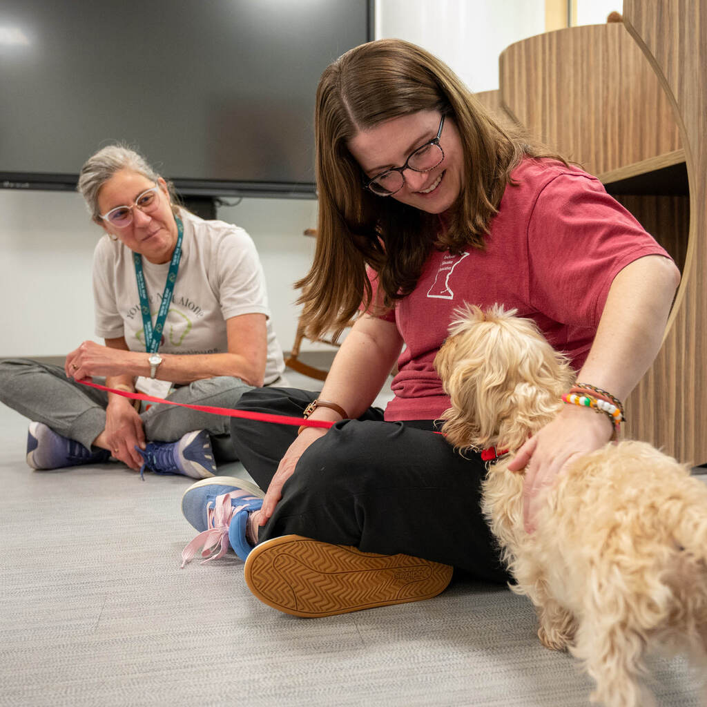 A teacher pets a small curly-haired dog on a leash. The woman holding the leash smiles at them.