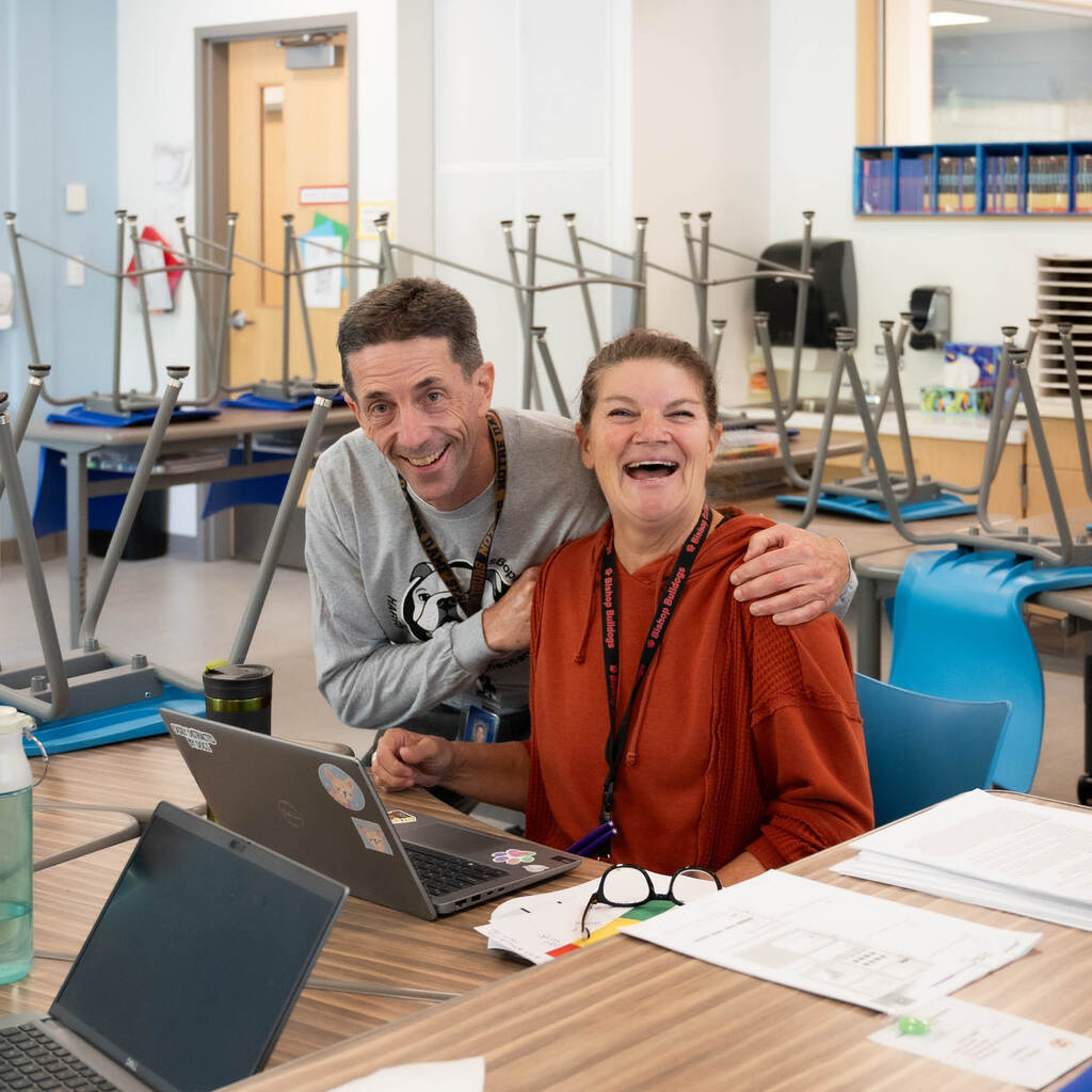 Two teachers smile at the camera in a classroom with the chair upsidedown on the tables.