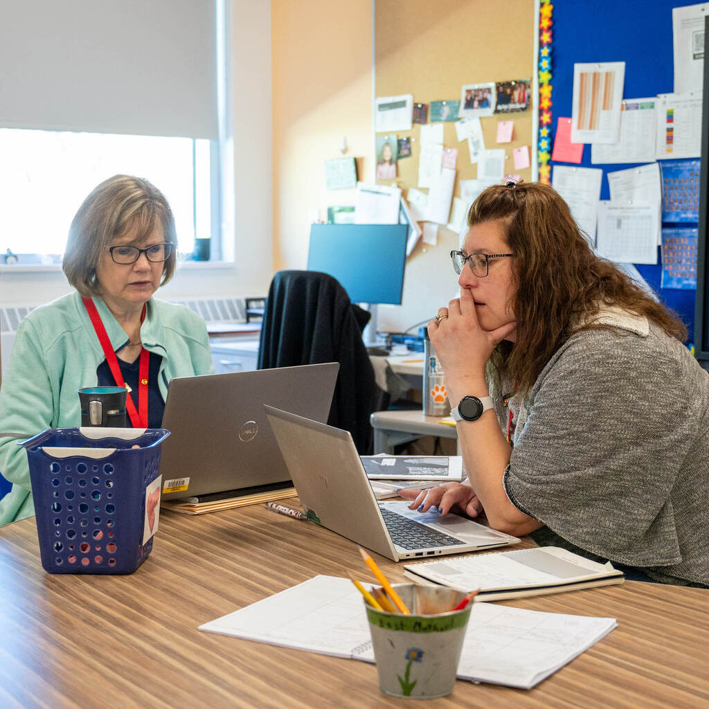 Two teachers at a table focus on their laptops.