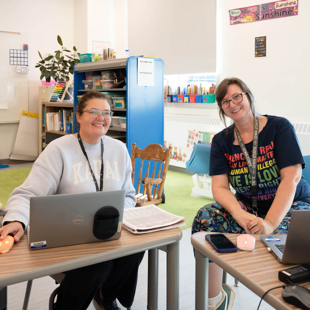 Two teachers smile at the camera while working on their laptops.