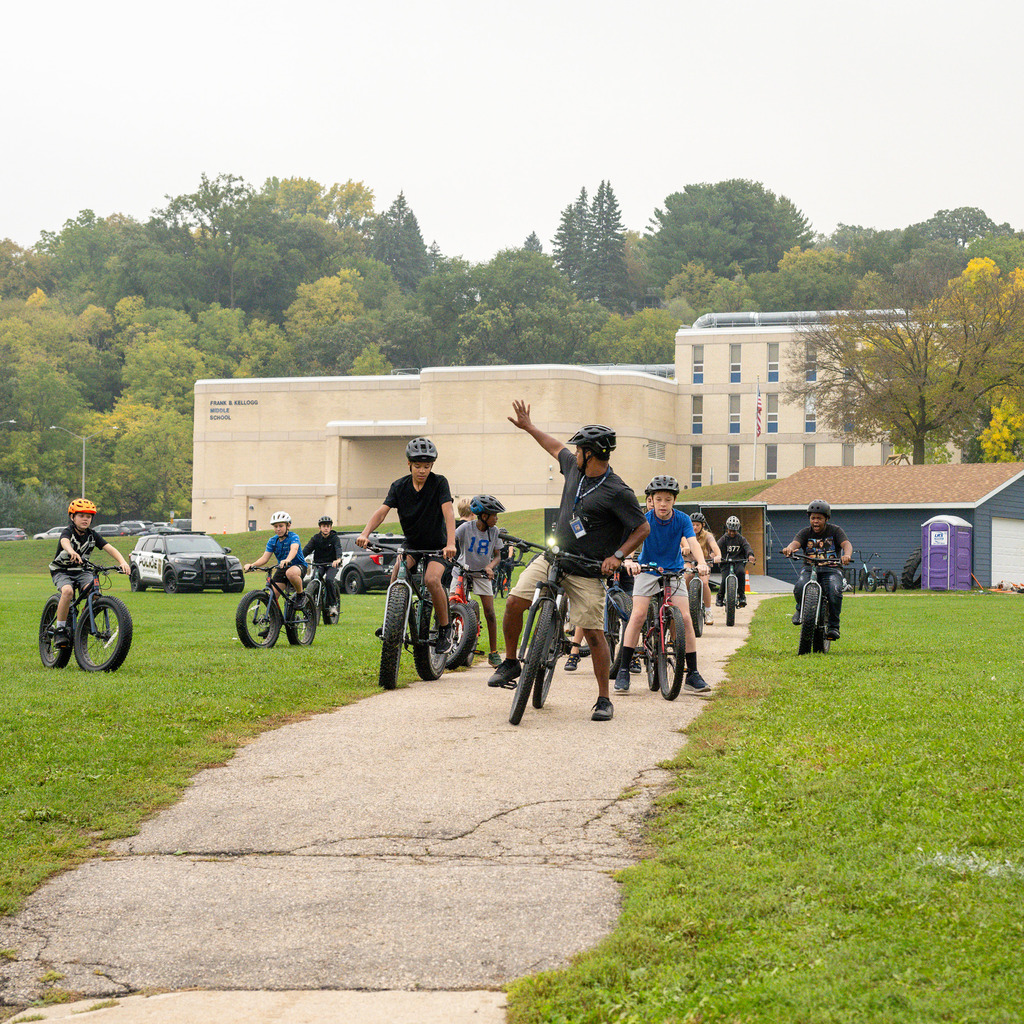 Out in front of a school, an instructor signals for a group of middle schoolers riding bikes to stop.