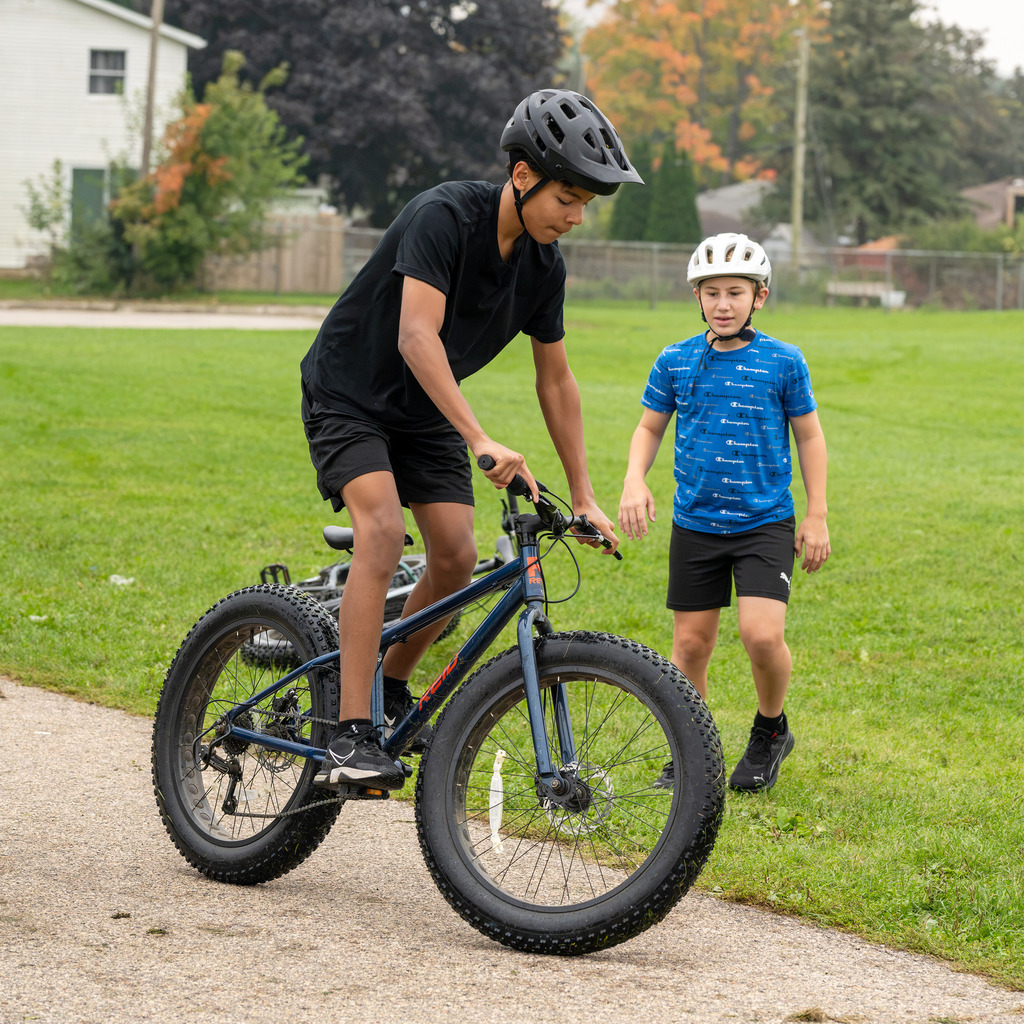 A boy tries out riding a flat-wheeled bike as another watches. Both are wearing helmets.