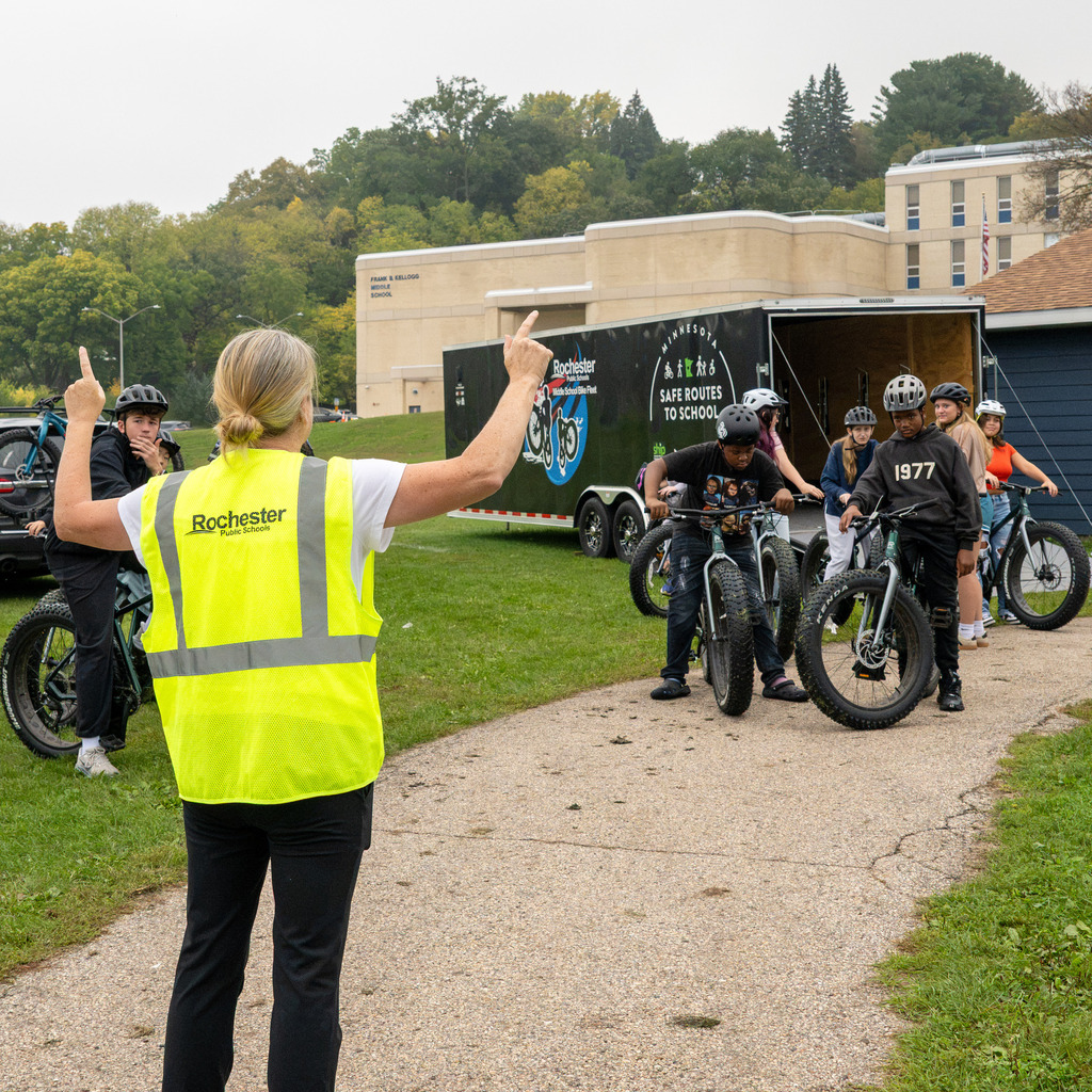 A woman in a neon safety vest gives riding and safety instructions to a group of students mounting bikes.