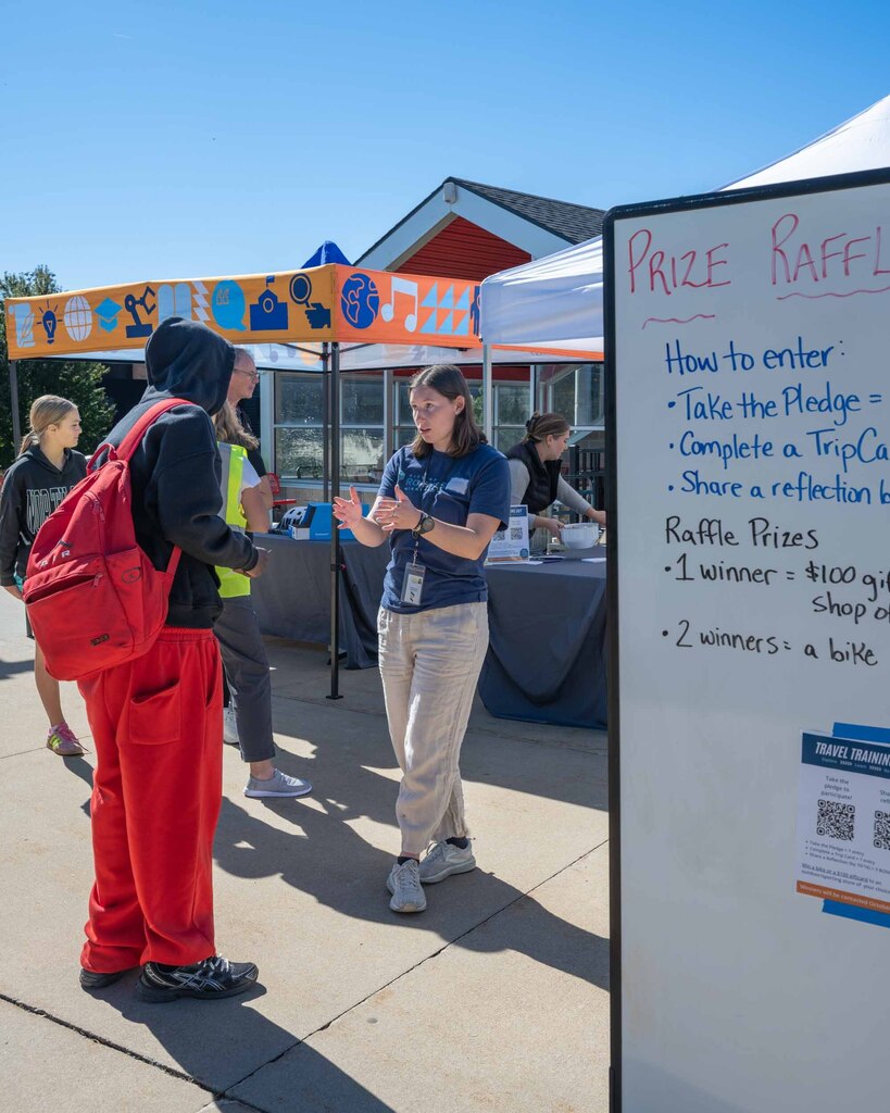 A woman wearing a lanyard talks to a student in a hoodie in front of a pop-up tent. A sing in the foreground explains how to enter a prize raffle.