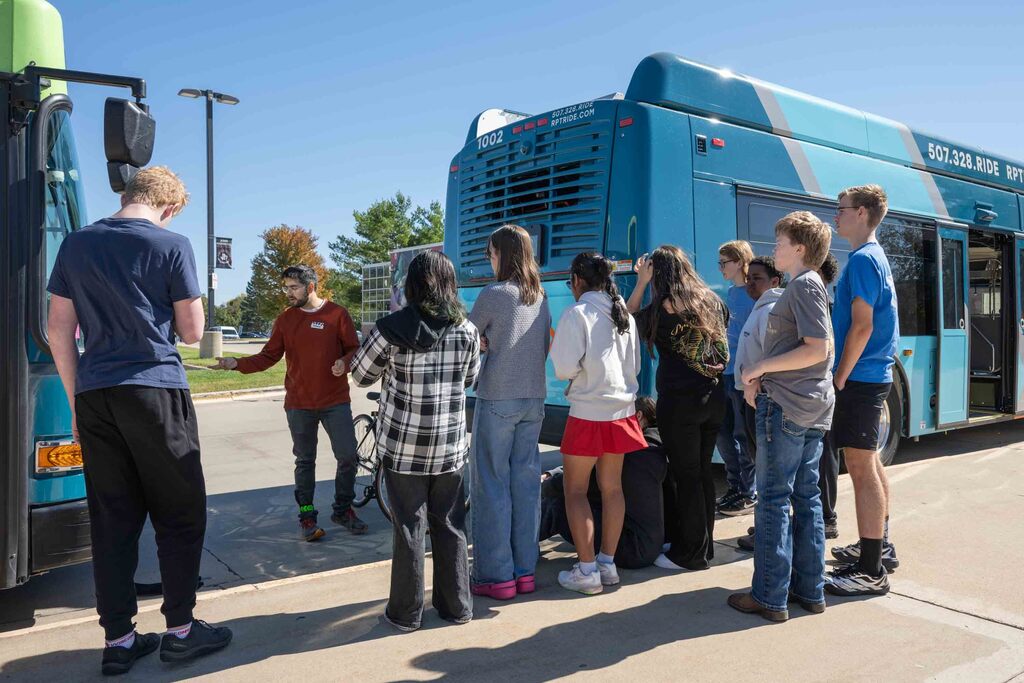 A group of high school students listen to an instructor as he shows them how to mount a bike on a city bus.
