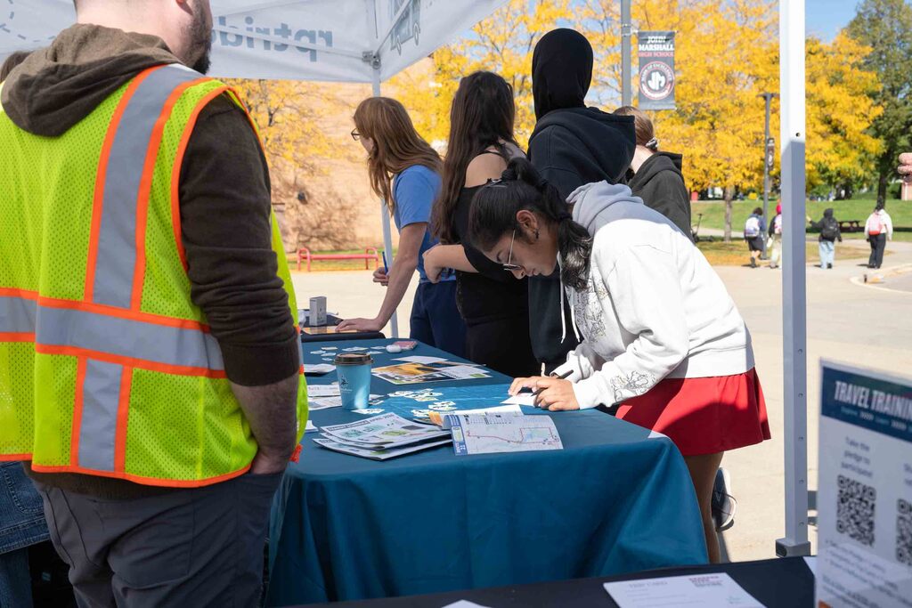 A student fills out a card at a tent table. A man in a safety vent stands in the foreground.