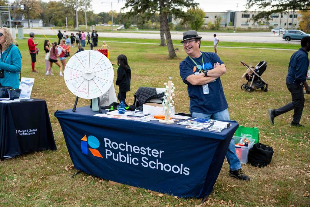 A man poses at a table that says Rochester Public Schools. There are give-aways and resources on the table.