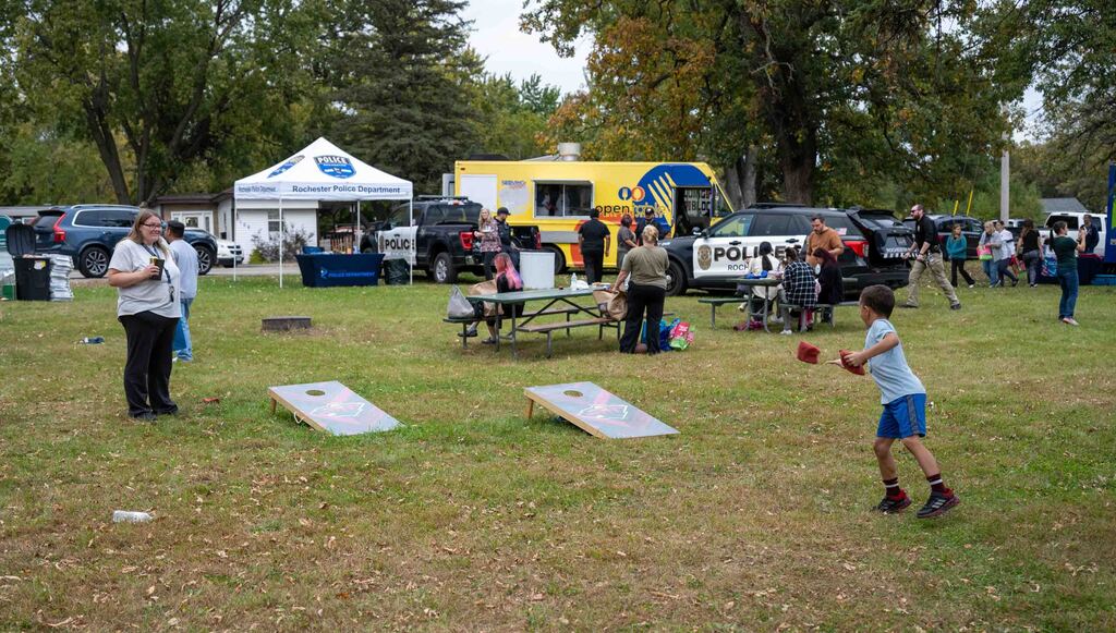 A kid throws a beanbag at a cornhole in a park.
