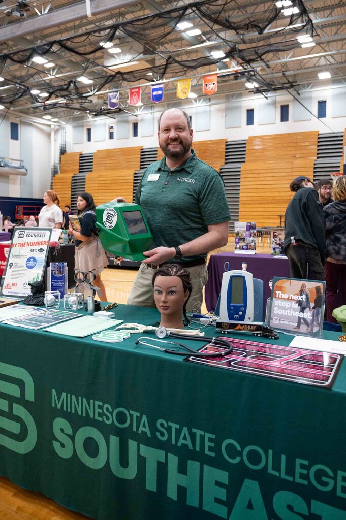 A man in a green polo golds a welding mask at a table presenting a college.