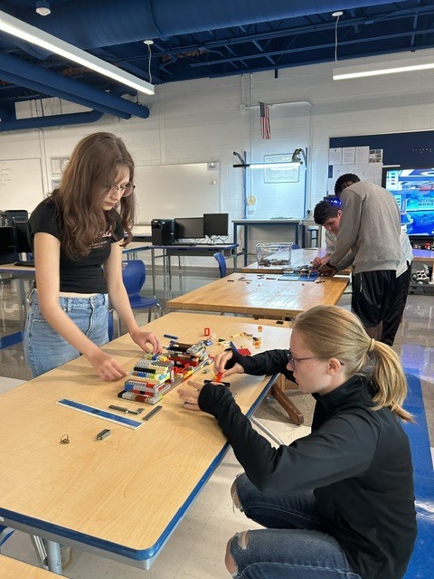 A classroom with white walls. Four people are working on a table with a variety of LEGO pieces.